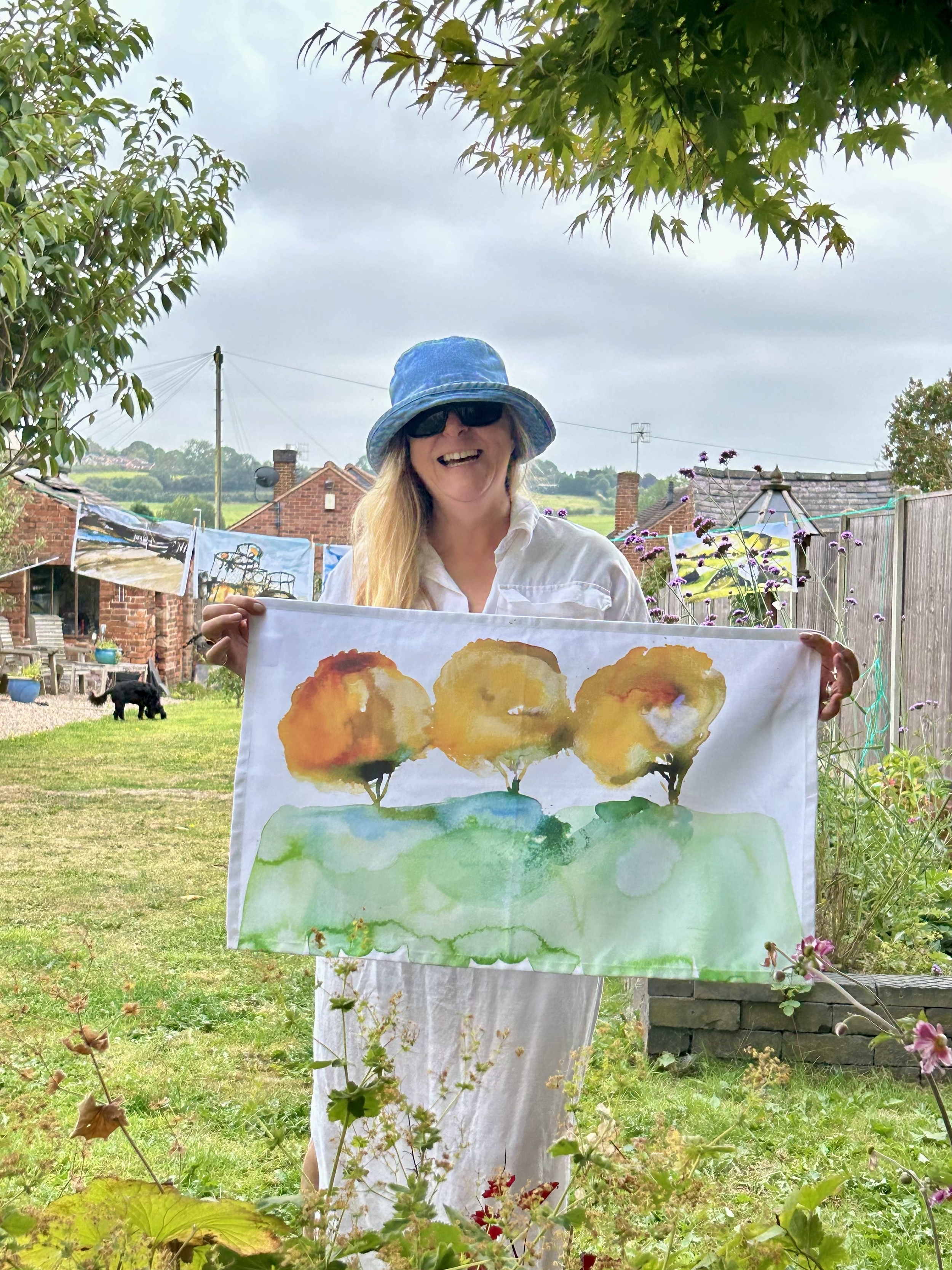 Woman wearing a blue bucket hat, sunglasses, and white dress stands in a garden holding a watercolor painting of three trees on a grassy hill. The sky is cloudy, and there are plants and flowers around her, with a brick house, clothesline with art, a