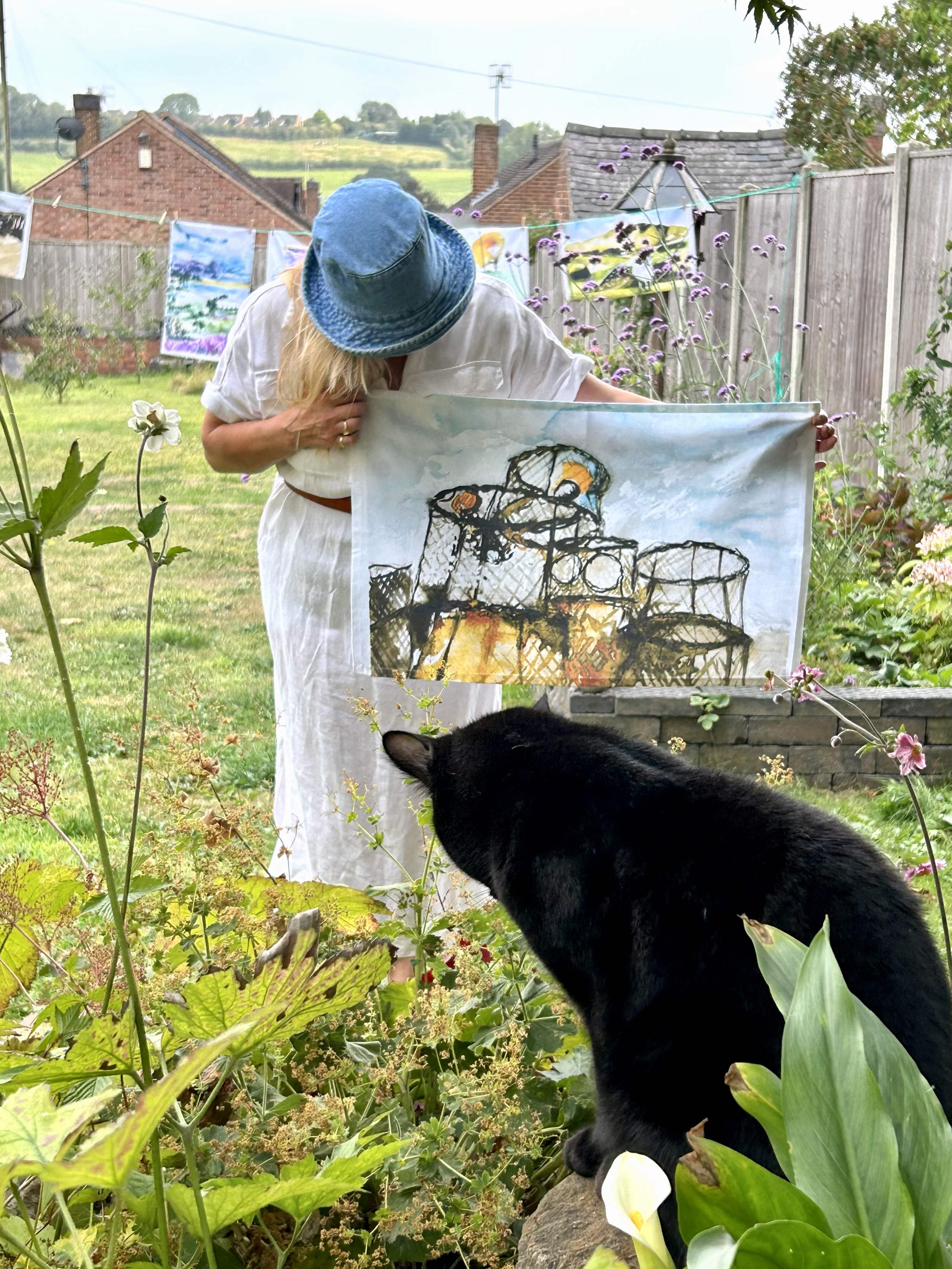 A woman with a blue hat and white dress holding a painting in a garden, with a black cat in the foreground sniffing plants, and clothespins with artwork hanging on a line in the background.