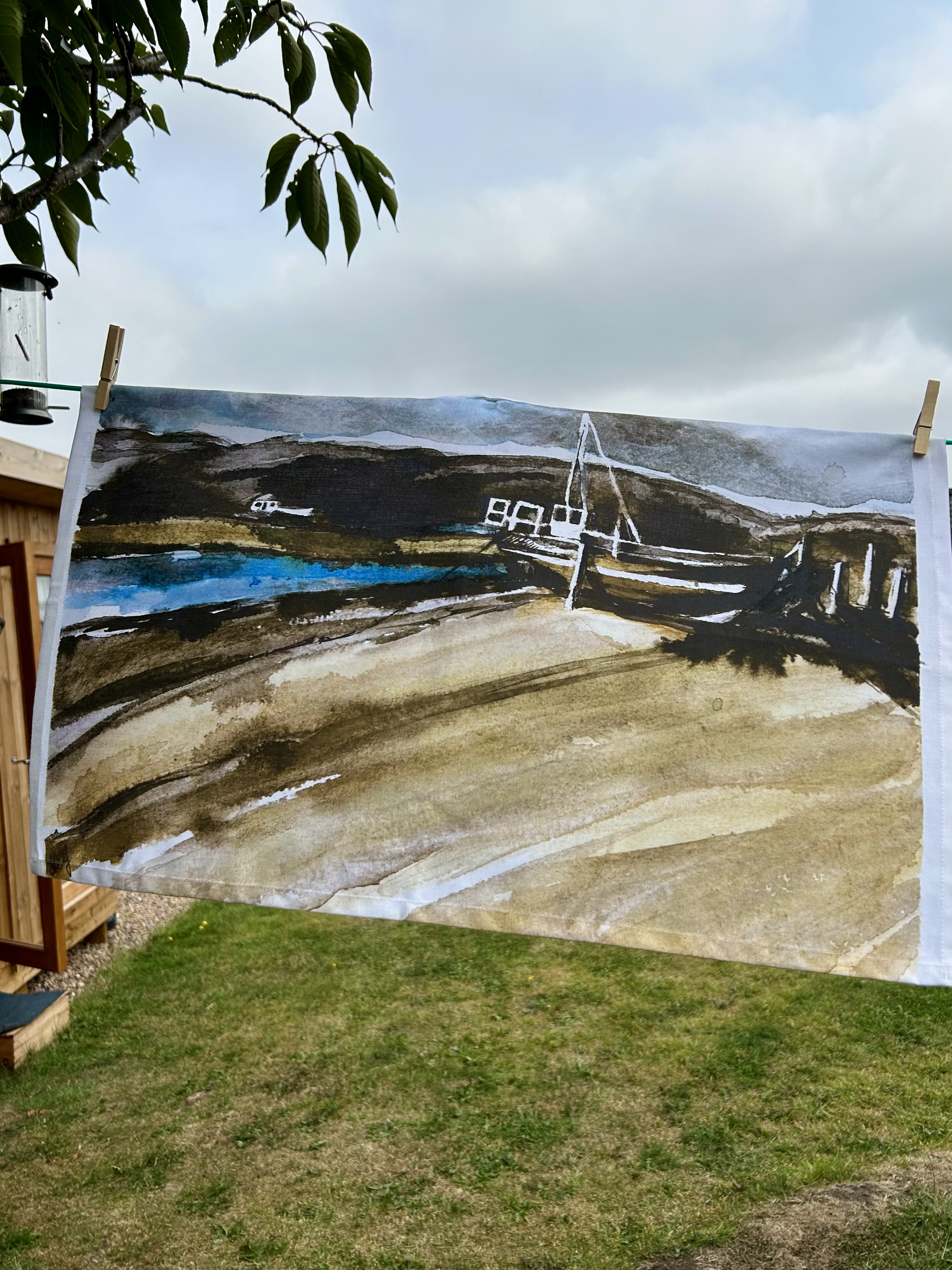 Watercolor painting of a landscape with mountains, water, and boats hanging outdoors on a clothesline with clothespins, with a grassy yard and cloudy sky in the background.
