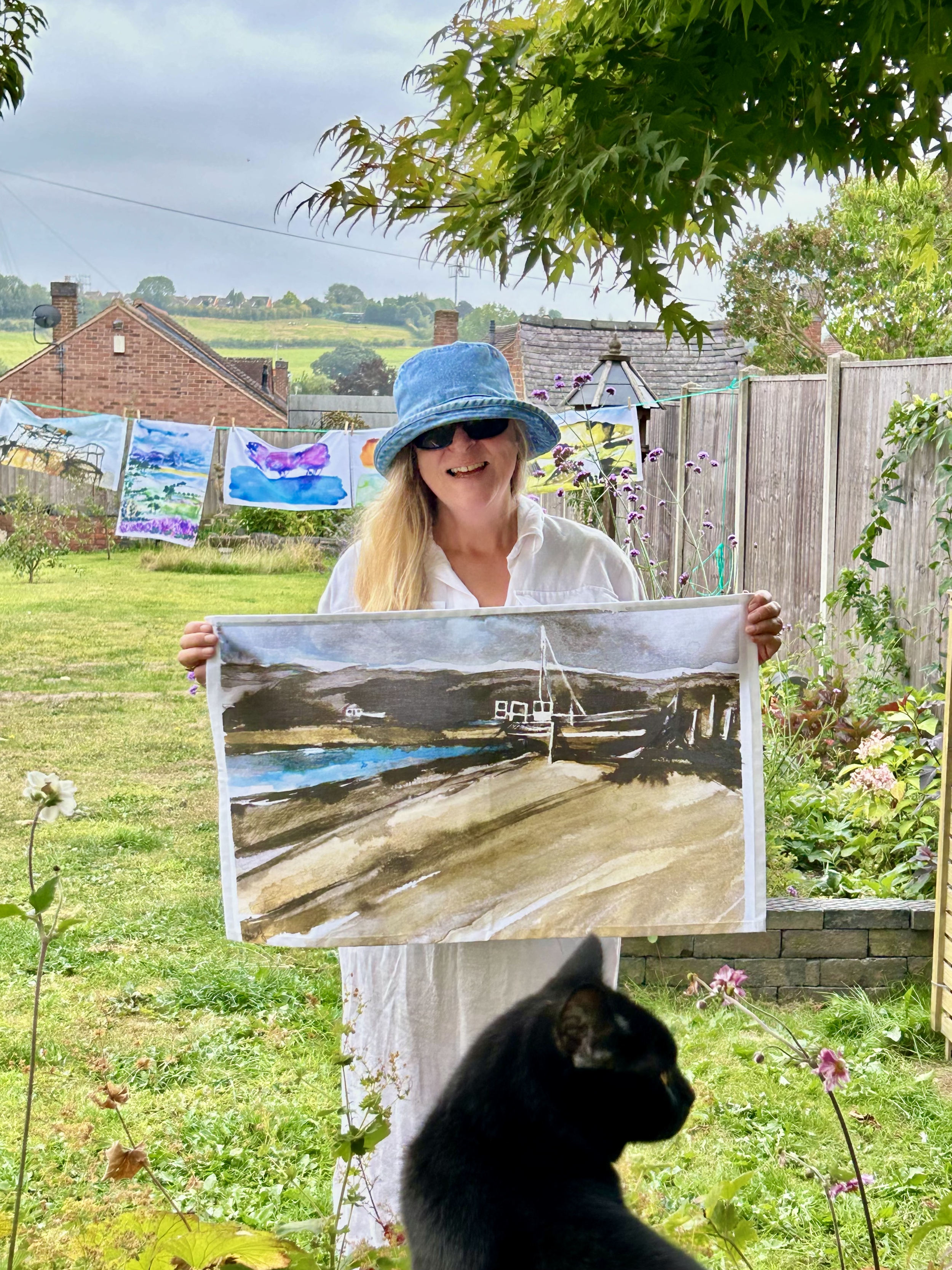 Woman with blonde hair, wearing a blue hat and sunglasses, holding a landscape watercolor painting outdoors in a garden. A black cat is in the foreground, and colorful artwork is hanging on a clothesline in the background.