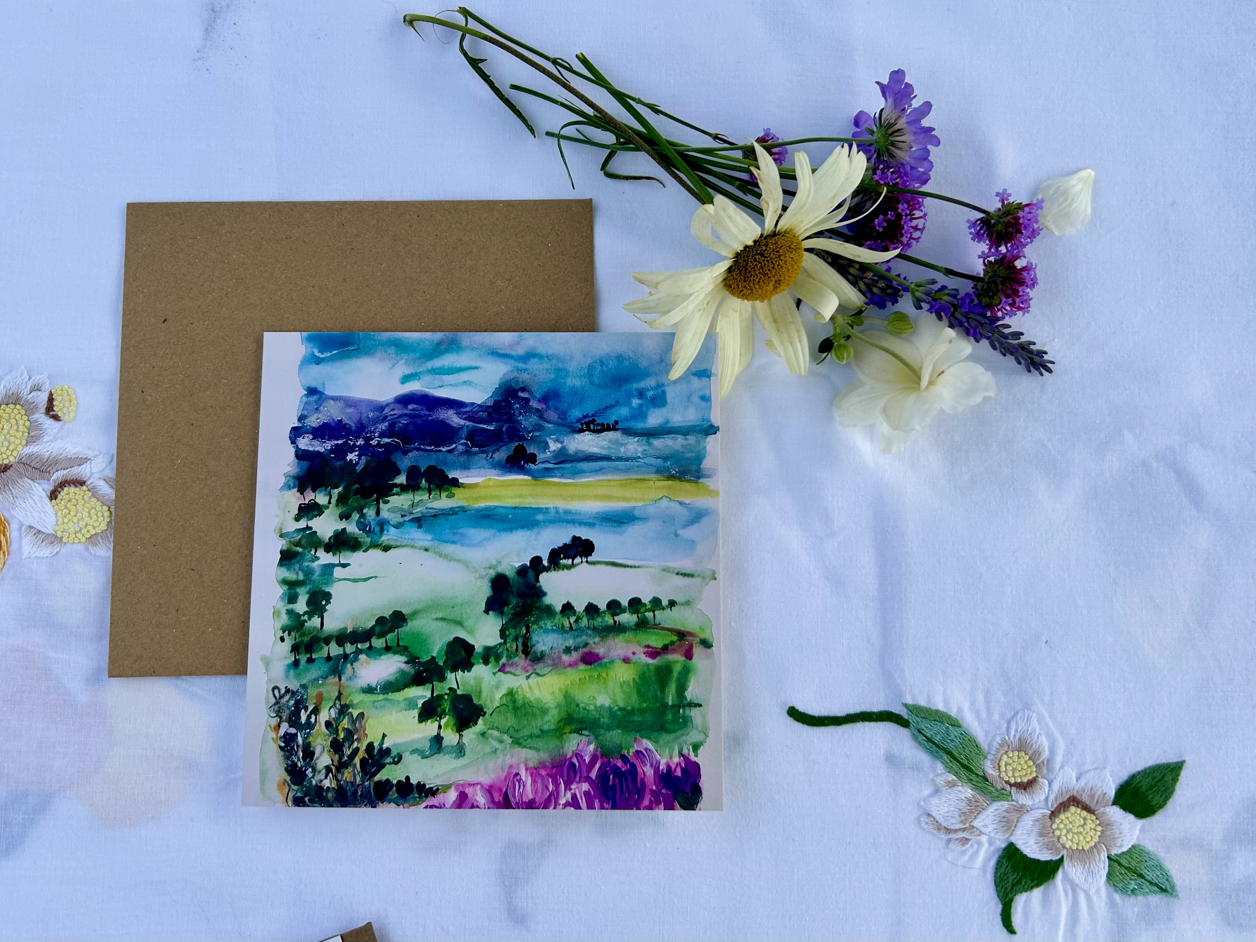 A watercolor painting of a landscape with hills, trees, and a cloudy sky, surrounded by a bunch of real white, purple, and yellow flowers on a white cloth.