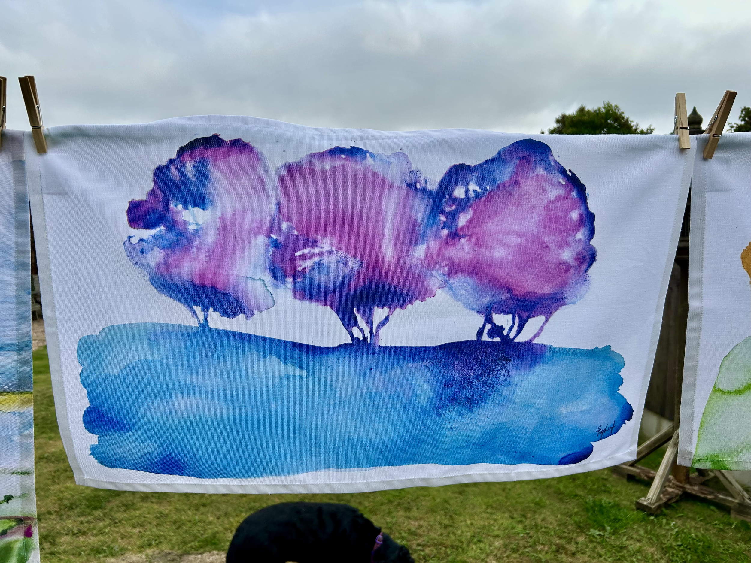 Watercolor painting of three purple trees on a grassy hill with a cloudy sky background.