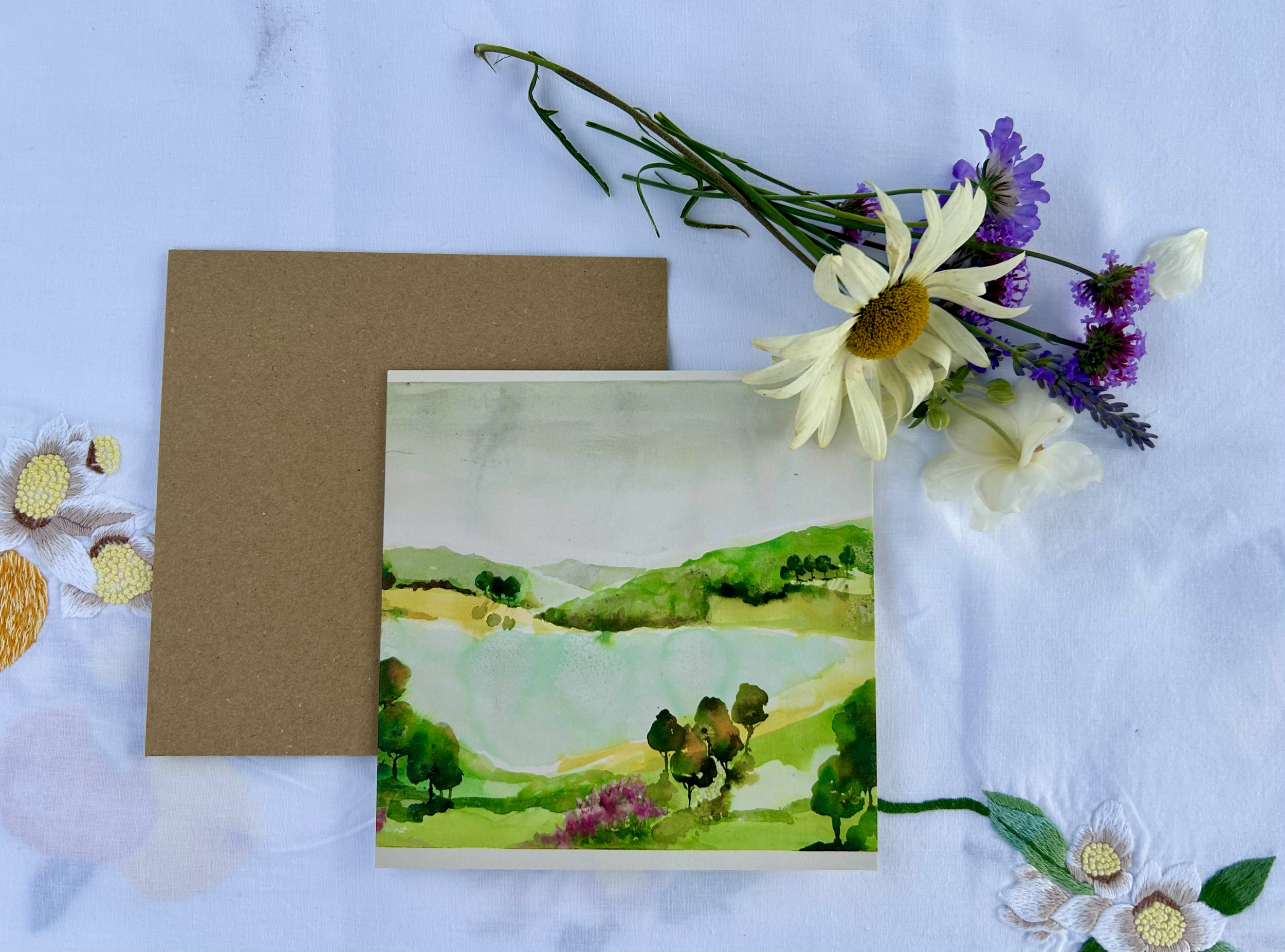 A handmade watercolor landscape painting of green hills, trees, a lake, and cloudy sky, placed on a white embroidered tablecloth with botanical decorations, a brown card, and a bouquet of white and purple flowers.