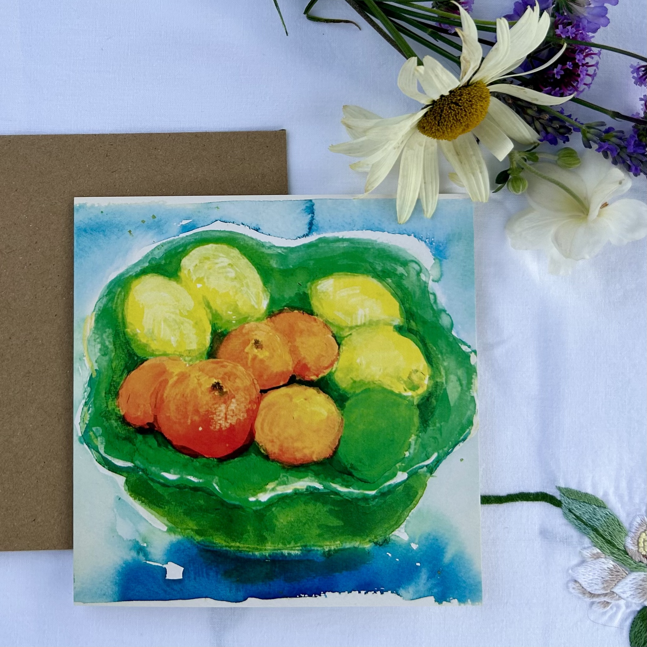 Watercolor painting of a bowl of assorted lemons and oranges, with a brown envelope and white flowers with purple accents nearby on a white tablecloth.