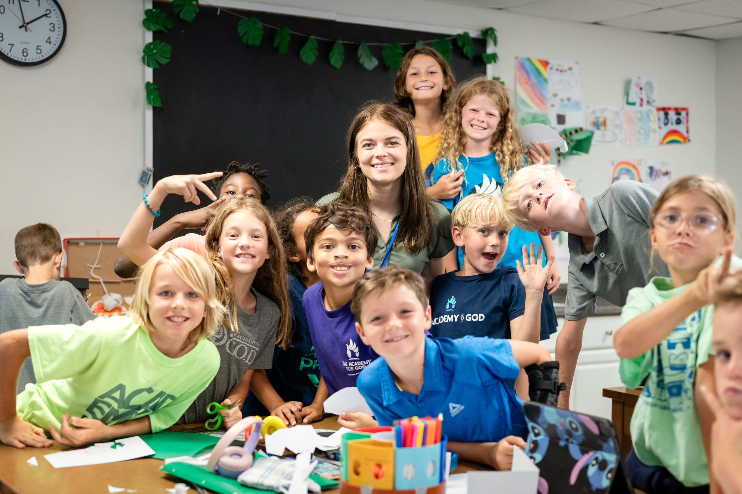 A group of smiling children gathered around a table covered with arts and crafts supplies in a classroom, with colorful artwork on the walls in the background.