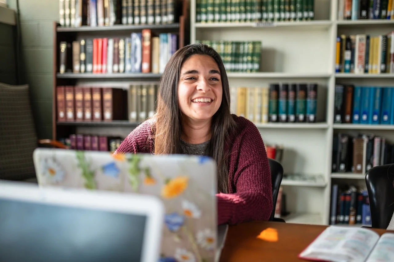 A woman with long dark hair smiling while sitting at a table in a library, with bookshelves filled with books in the background.