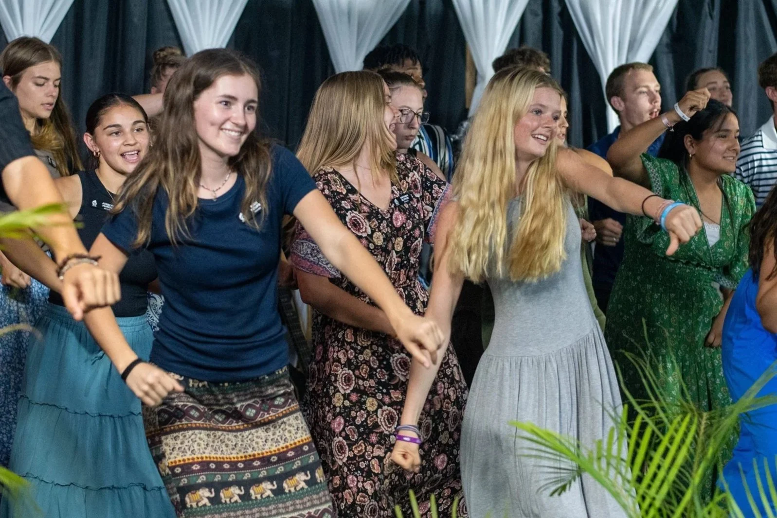 Group of young people dancing and smiling in a decorated indoor party with white and dark blue curtains.