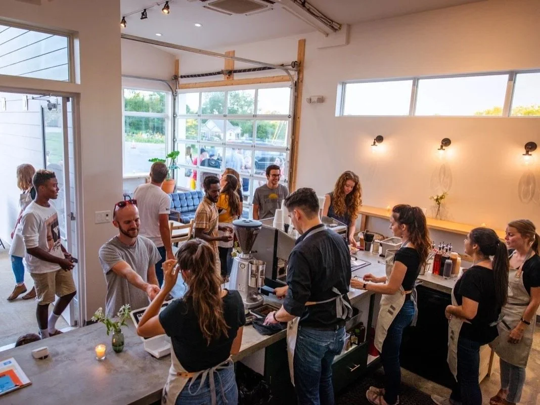 People inside a coffee shop or cafe, some customers ordering at the counter and others waiting or sitting near the window, with large windows letting in natural light.