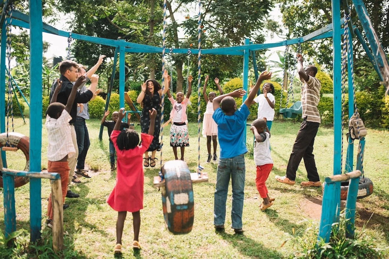 A group of children and an adult are playing and jumping on swings and tires in a playground outdoors, surrounded by trees and greenery.