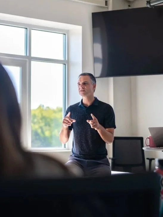 Man giving a presentation in a bright room with a large window and a big screen.