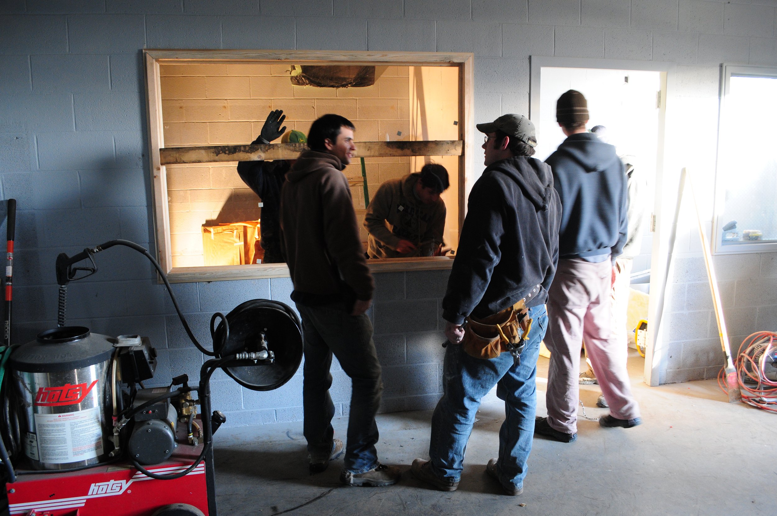 A group of people working together inside a garage or workshop, with a window opening to the other room, various equipment, and tools around.