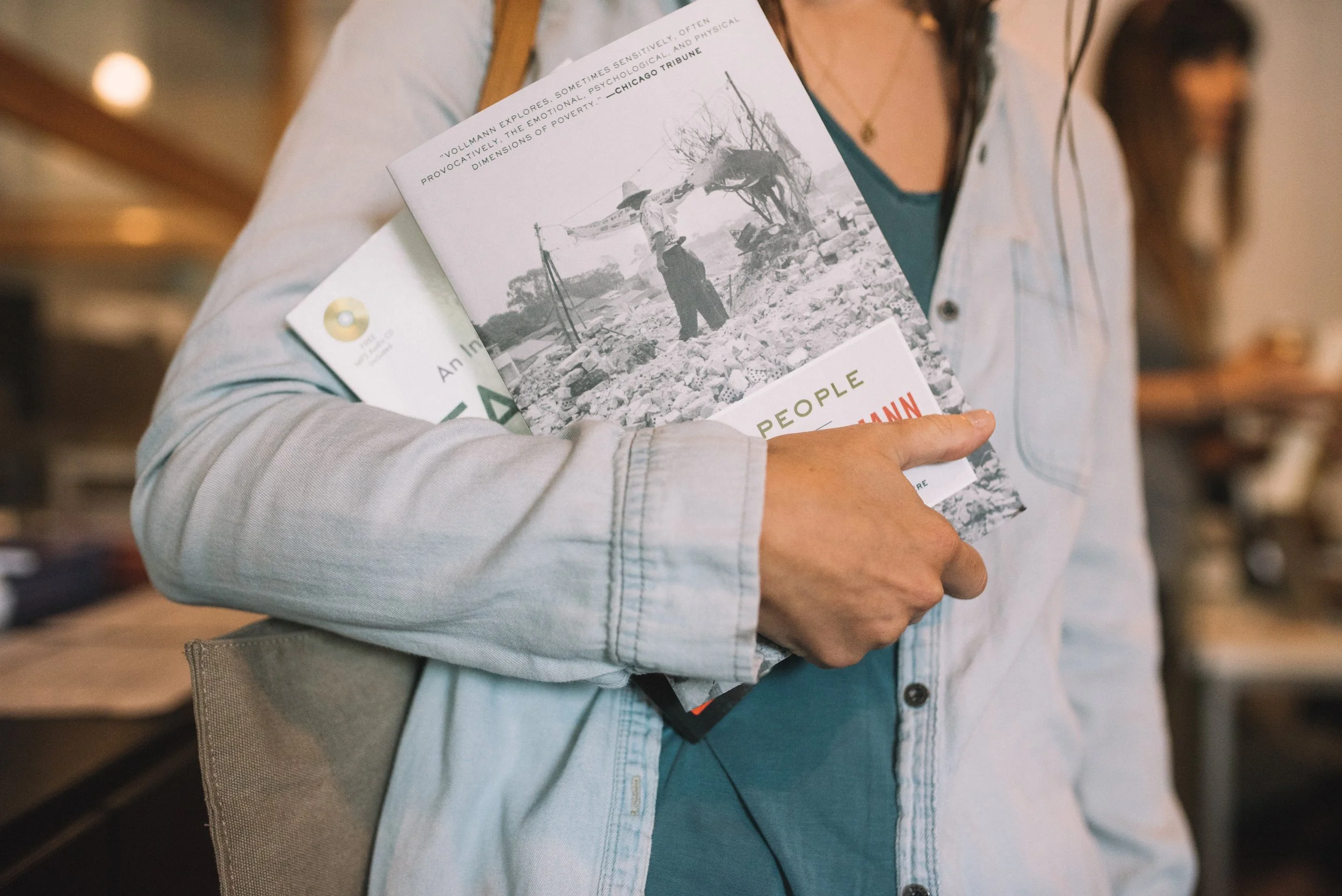 A person holding a brochure or booklet with a black-and-white photo of a man standing near trees, holding an umbrella. The person is wearing a light-colored denim jacket and a dark teal shirt underneath.