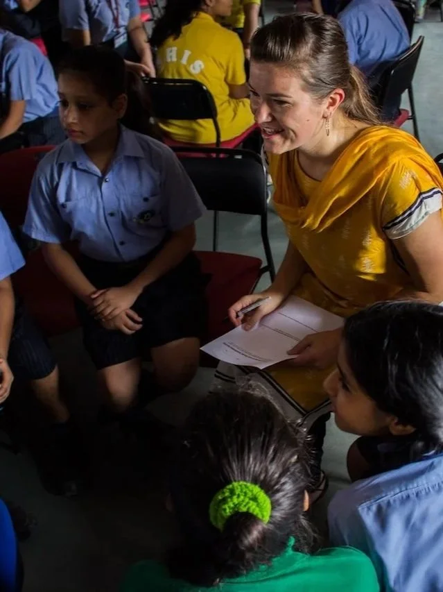 A woman in a yellow traditional outfit is smiling and talking to children in school uniforms, seated in a circle, in a classroom or school setting.