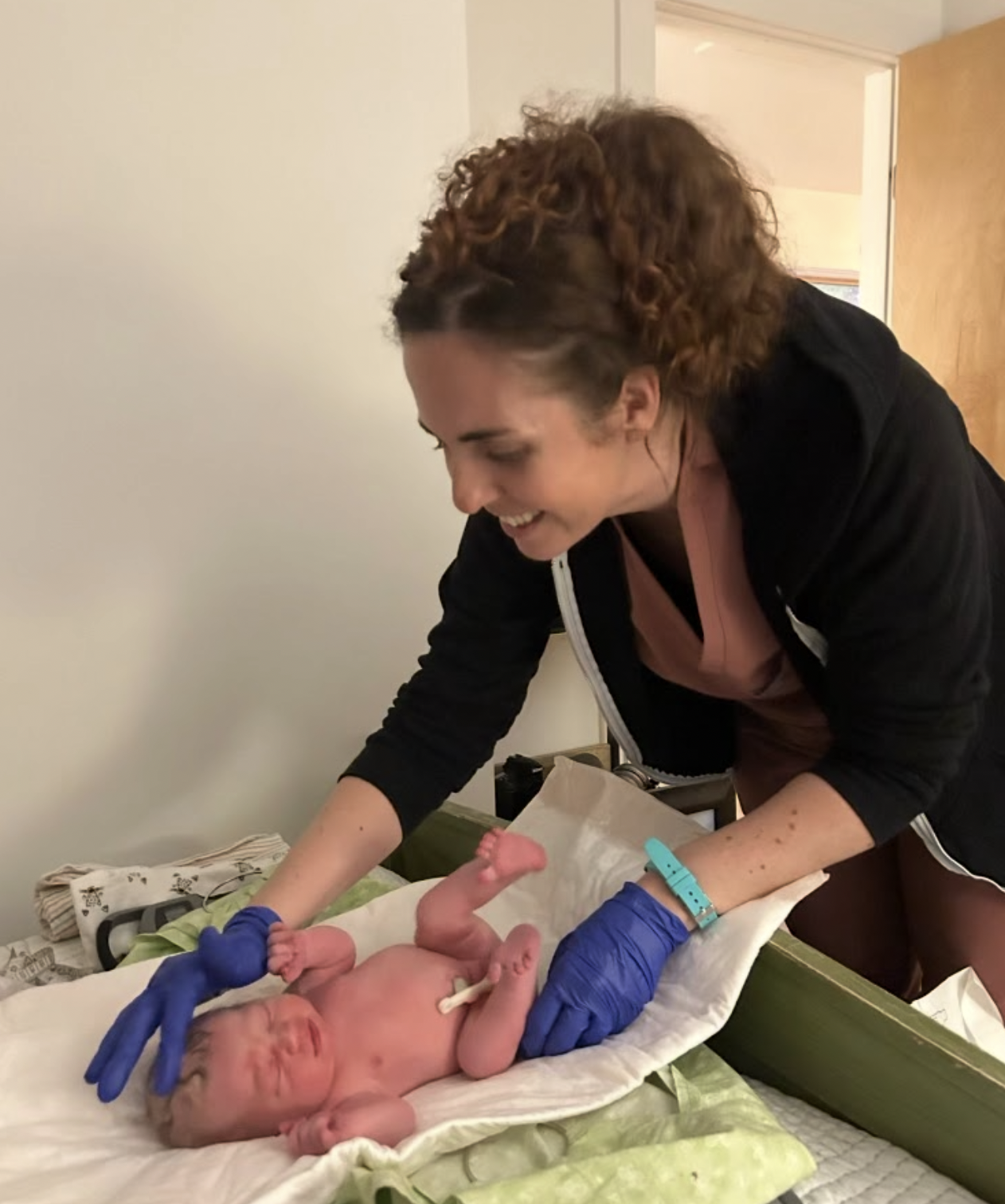 A woman with curly hair leaning over a hospital bassinet, smiling at a newborn baby wrapped in blue gloves. The baby has a band around one ankle and appears to be just born.