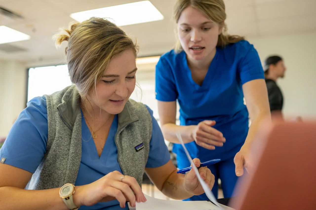 Two female healthcare workers in blue scrubs discussing paperwork indoors.