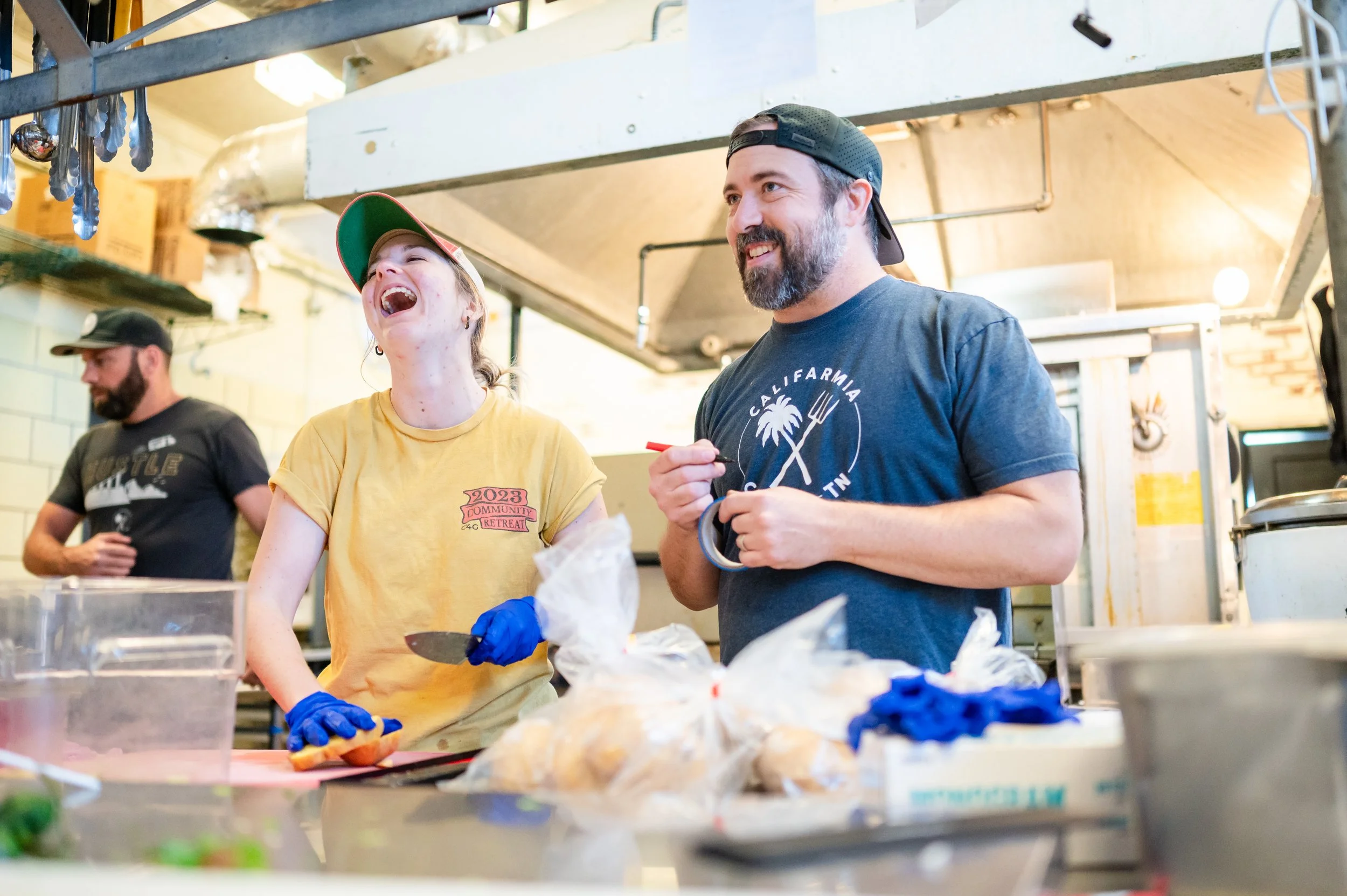 Two people, a woman and a man, smiling and laughing while working in a kitchen or food prep area. The woman wears a yellow shirt and a cap, and the man wears a navy t-shirt and a backward cap. They are preparing food, possibly at a community or casual event, with various kitchen supplies and ingredients on the counter in front of them.