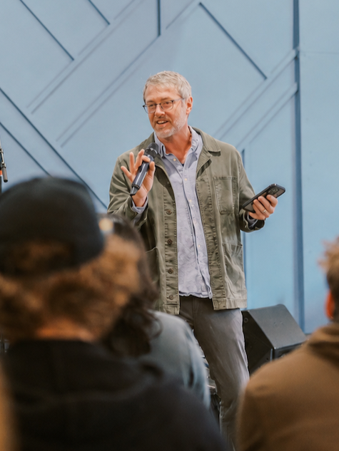 A man with gray hair, glasses, wearing a green jacket, speaking into a microphone during a presentation or speech.