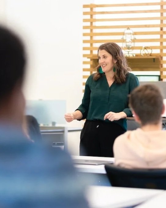 A woman standing and speaking to a group in a modern classroom or office setting, with a wooden slat wall in the background.