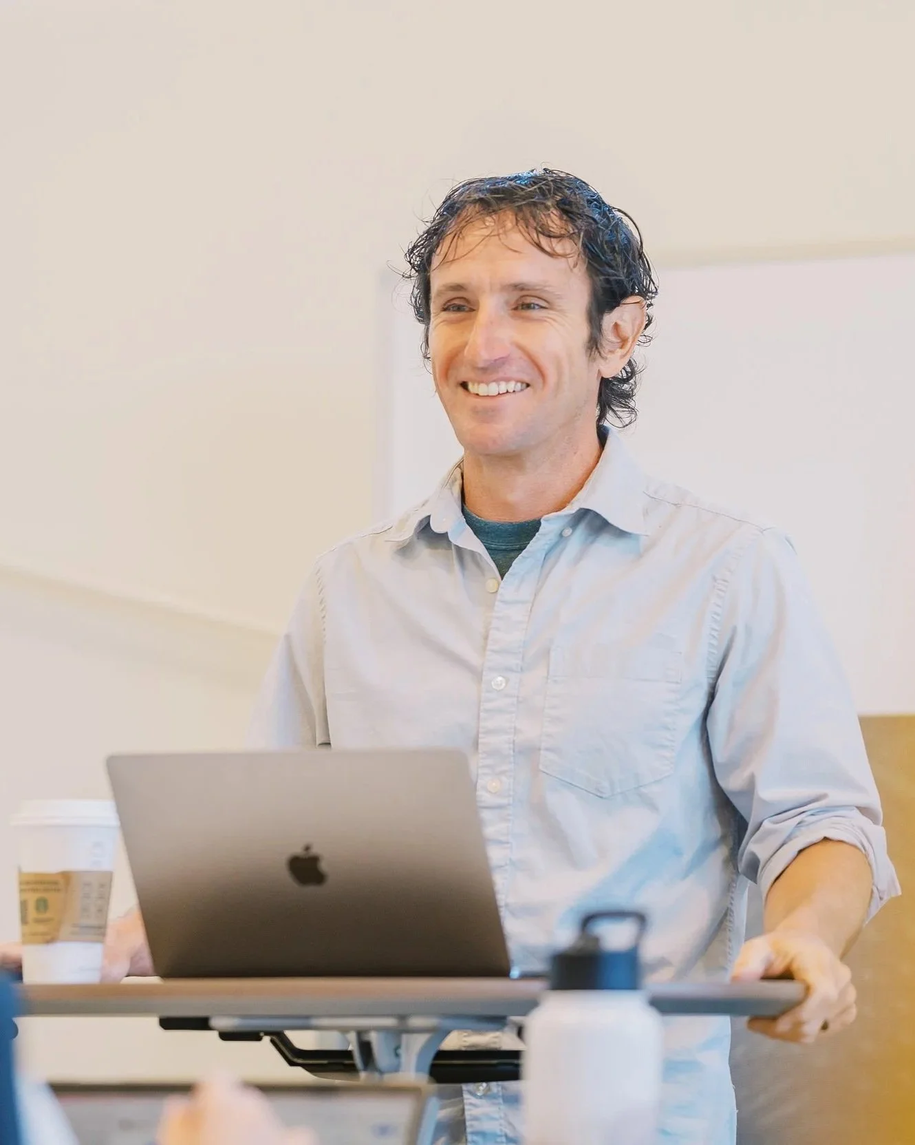 A man with curly brown hair smiling while working on a laptop at a desk in a bright room.