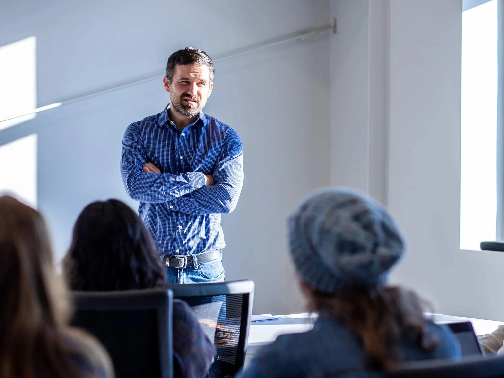 A man with dark hair and a beard wearing a blue checkered shirt stands with crossed arms in front of a group of people in a classroom or conference room. He appears to be speaking or presenting to an audience, with sunlight streaming in through windows on the right side of the image.