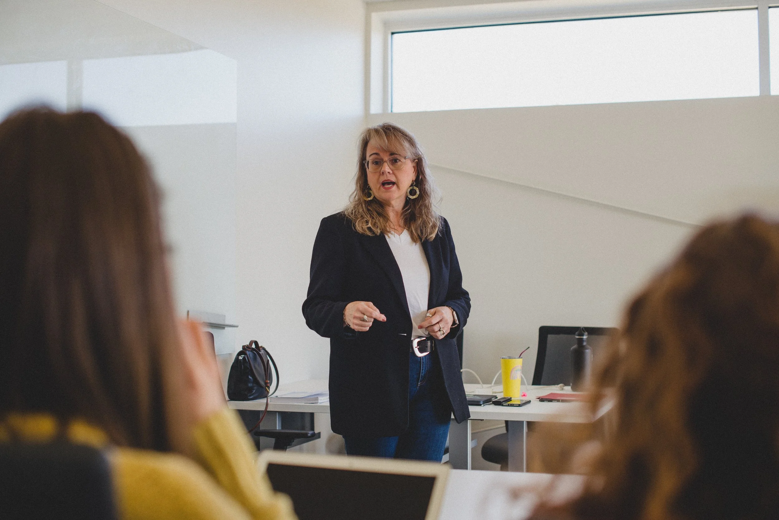 A woman with glasses and earrings speaking to a group in a modern classroom or conference room, with people seated at desks in the foreground.