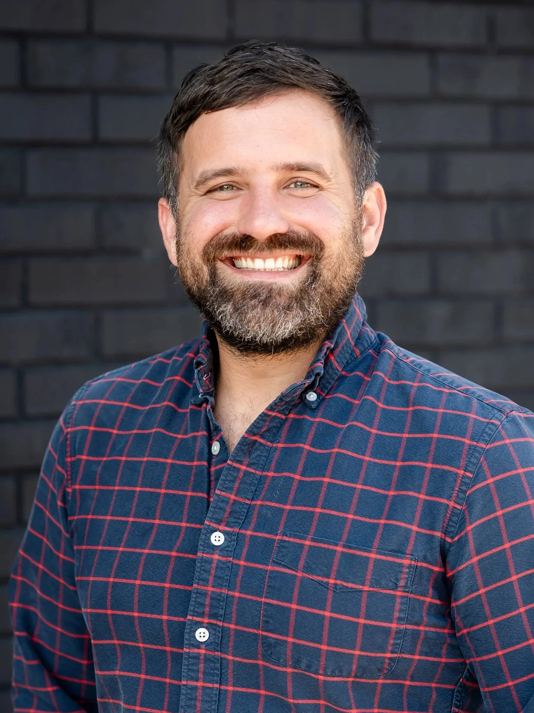 A man with a beard and short hair smiling, holding a green bottle, at a gathering with others, in a bright room with large window