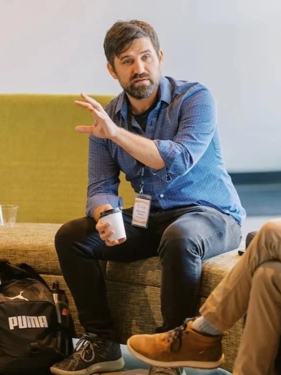 A man with a beard and dark hair, wearing a blue checkered shirt and black pants, sitting on a bench, gesturing with his right hand while holding a coffee cup in his left hand. There is a backpack and a person wearing tan pants and brown boots next to him.