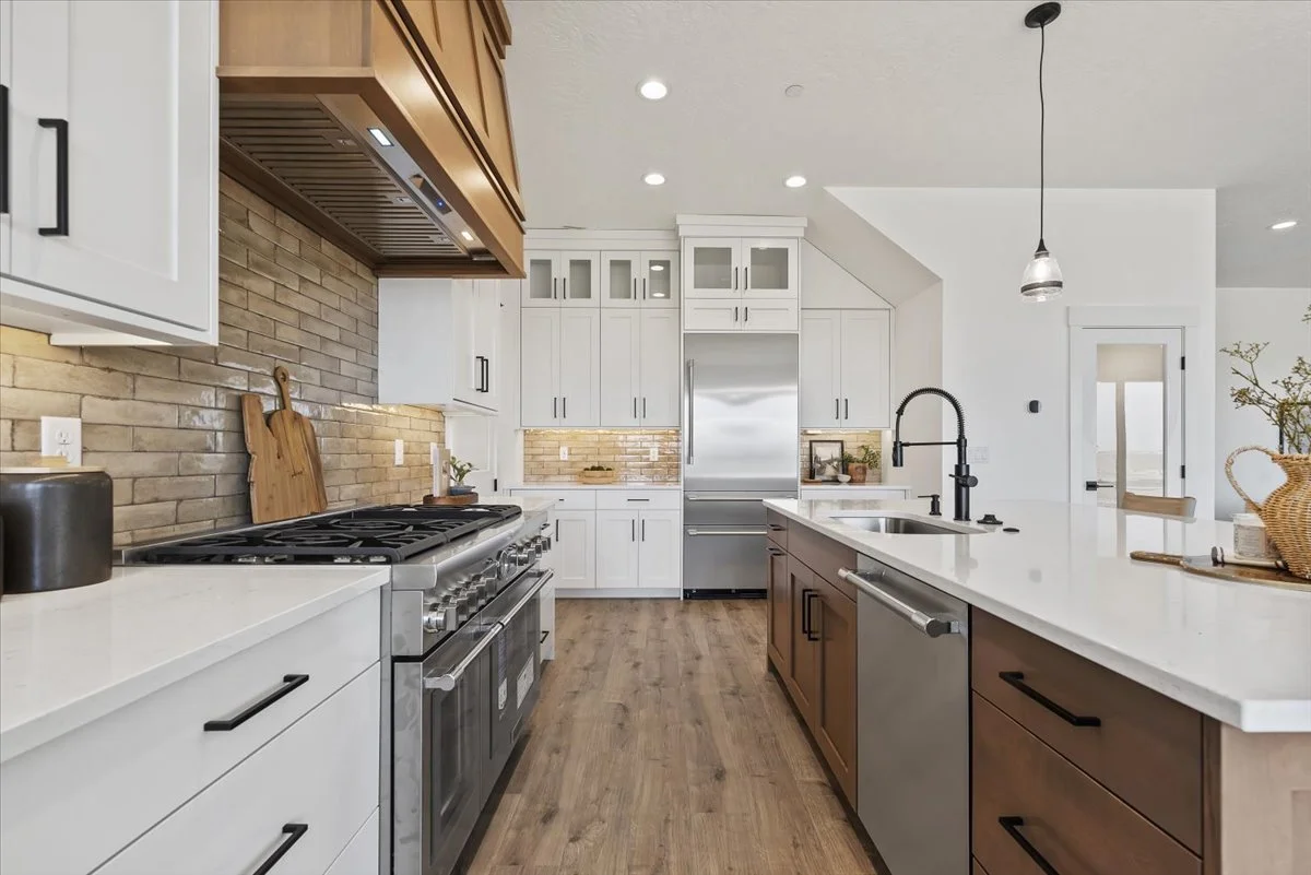Modern open-concept kitchen with wooden cabinets, stainless steel appliances, a large white island with a faucet, and a living area with a gray sectional sofa and large windows.