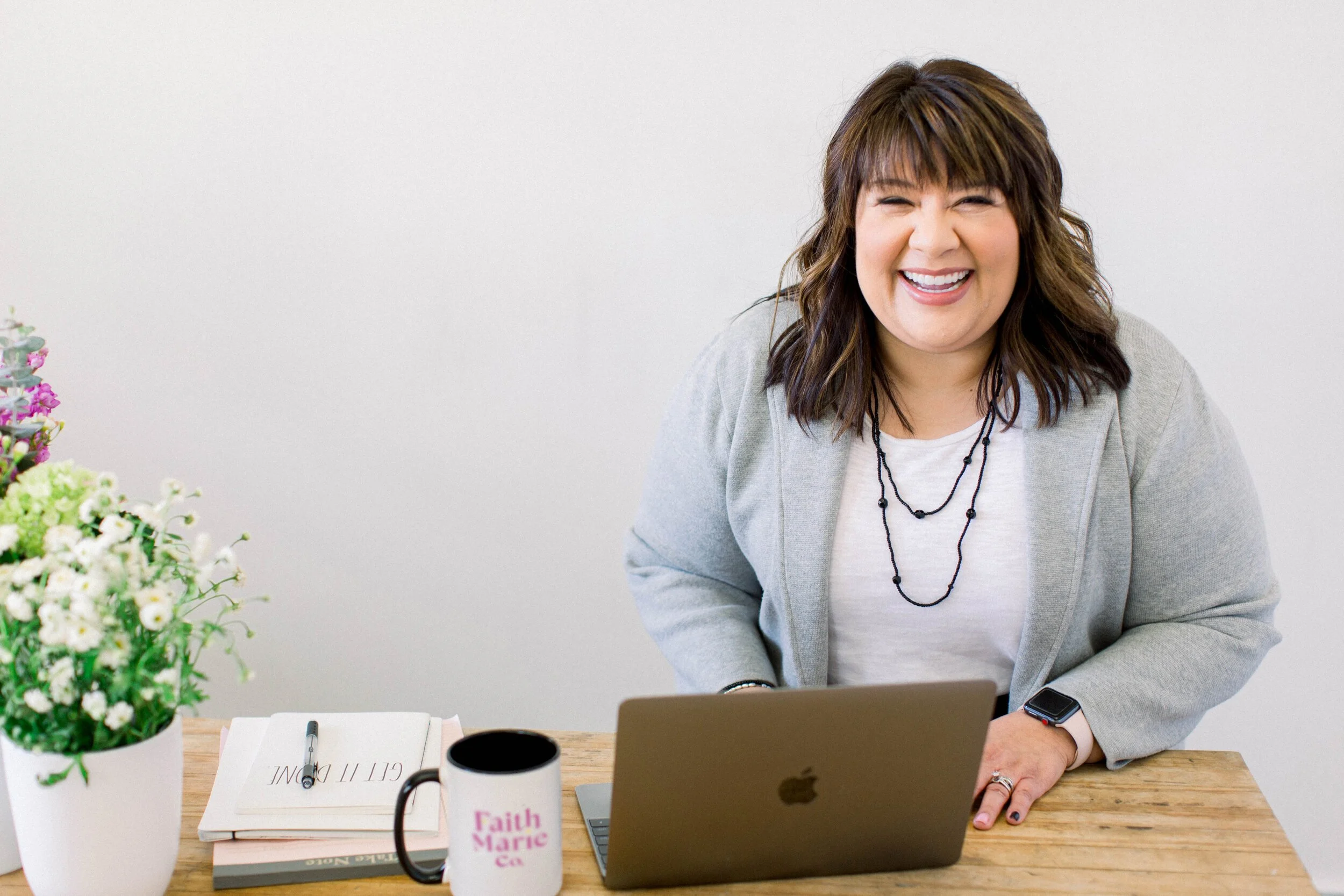 A woman with shoulder-length brown hair, smiling, sitting at a desk with a laptop. She is wearing a light gray blazer, white t-shirt, black necklace, and smartwatch. On the desk are a notebook, pen, coffee mug with pink text, and a vase of flowers. The background is plain and white.