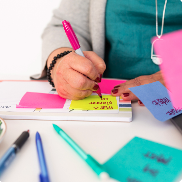 Person writing on a pink sticky note with a pink marker, surrounded by colorful pens and notes on a white desk.