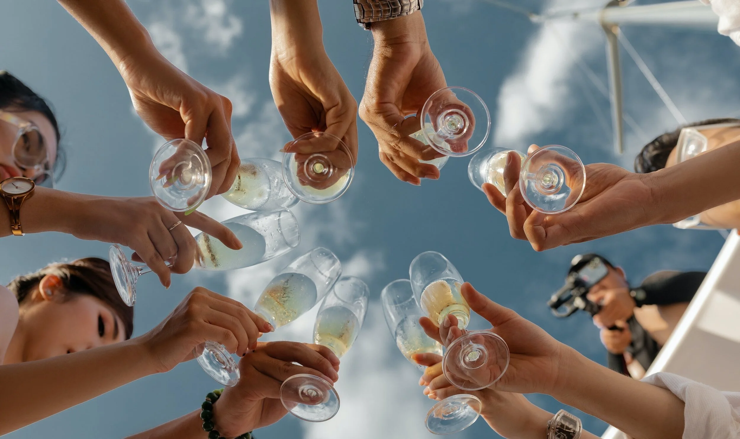 People clinking glasses of champagne in a toast outdoors against a bright blue sky with clouds.