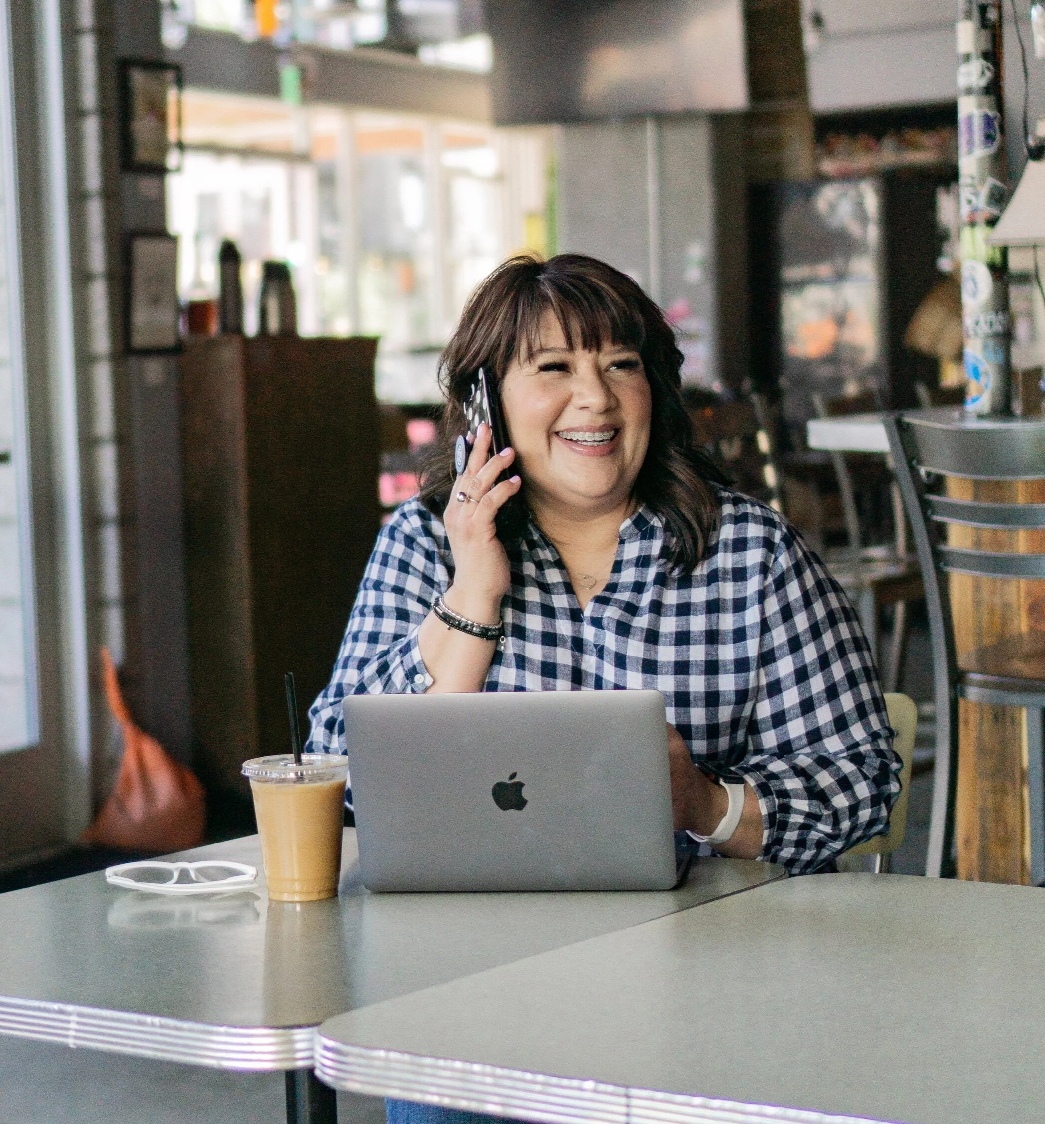 A woman with dark hair, wearing a blue and white checkered shirt, is talking on a cell phone and smiling. She is sitting at a table with a silver MacBook laptop, a yellow cold drink, and sunglasses.