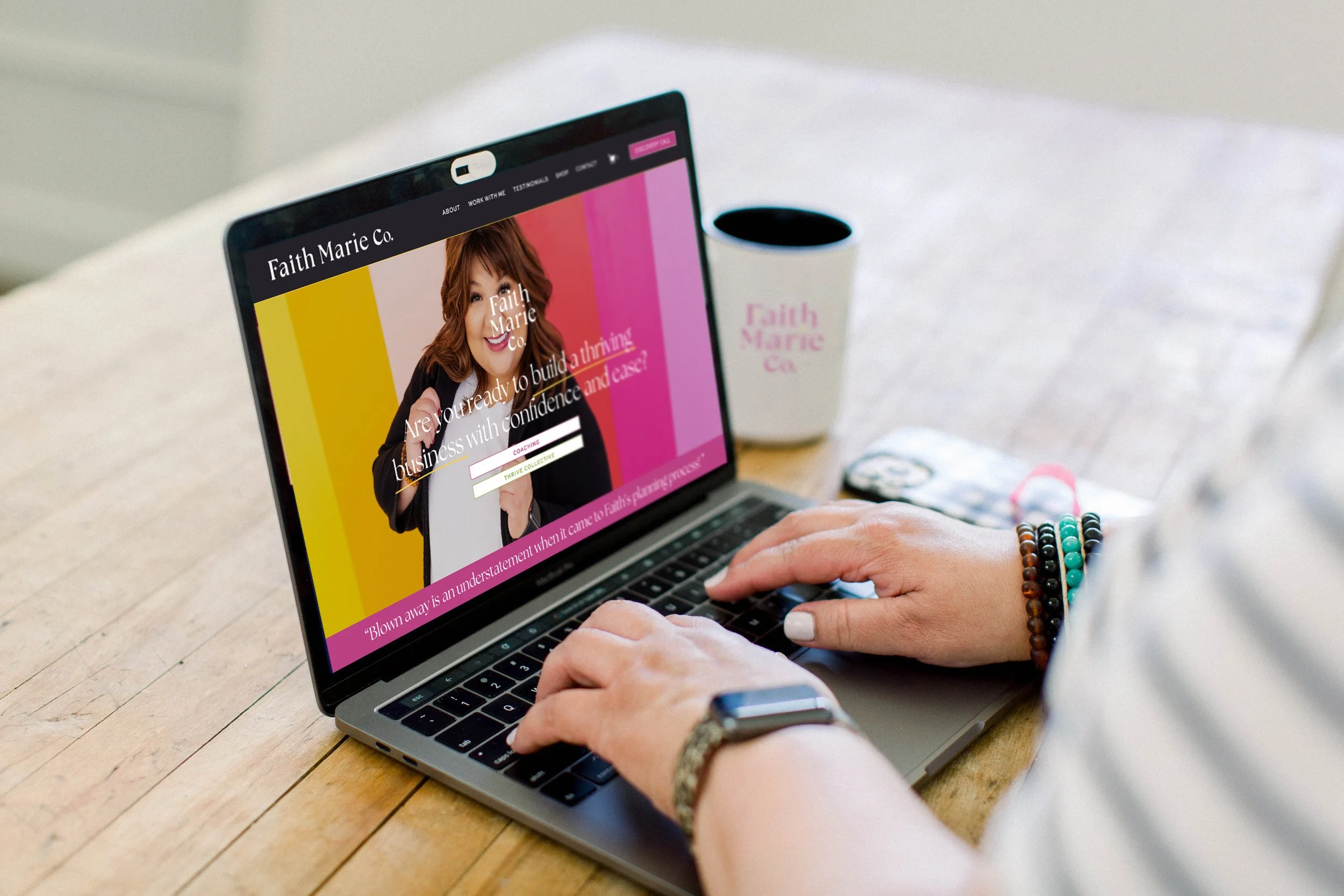 Person using a laptop with a website for Faith Marie Co. open on the screen, sitting at a wooden table with a branded coffee mug and a snack.