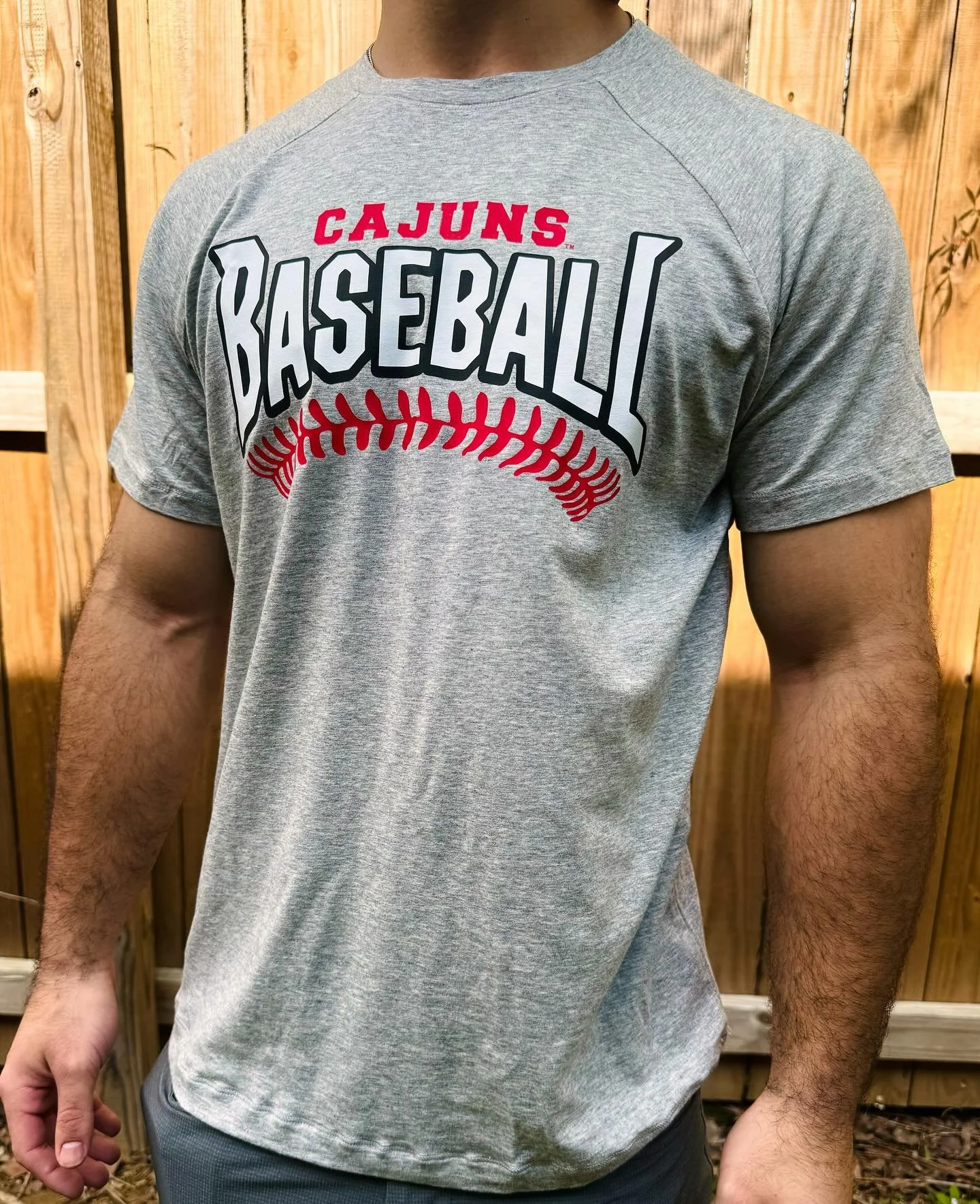 A person wearing a gray T-shirt with 'Cajuns Baseball' written on it, standing in front of a wooden fence.