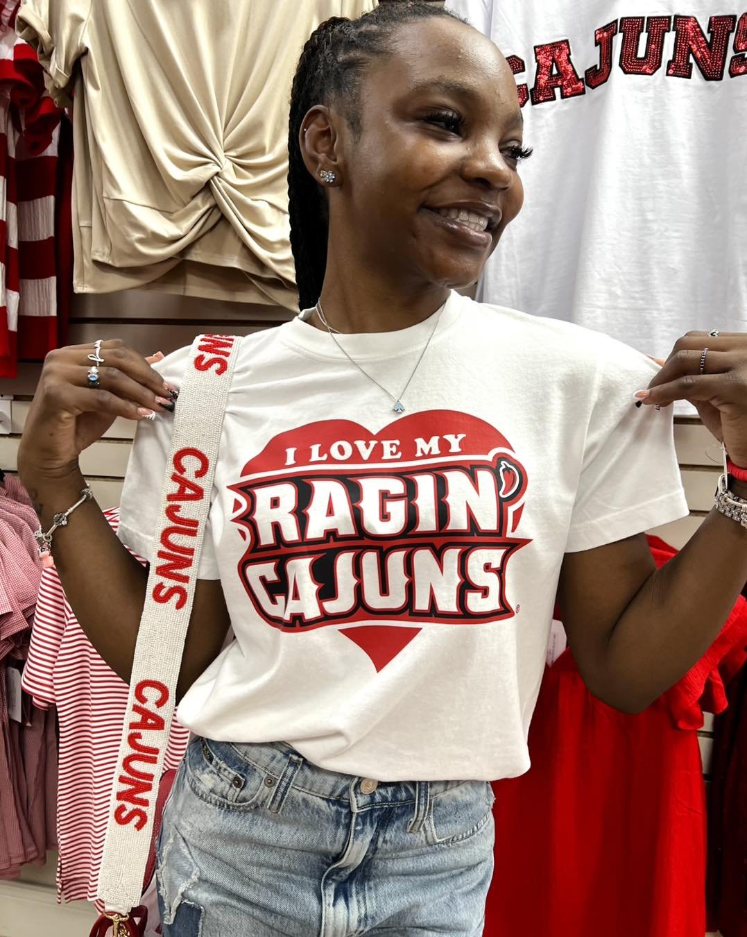 A woman wearing a white t-shirt with red and black text that says "I love my Ragin Cajuns," standing in front of a rack of red and white clothing. She is smiling and pointing to her shirt, with her hair styled in braids and wearing jewelry. She is in