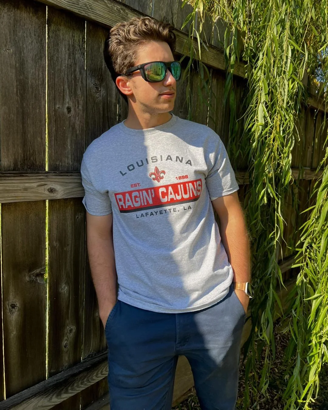 Young man wearing sunglasses and a grey T-shirt with 'Ragin' Cajuns' text, standing by a wooden fence with greenery nearby.
