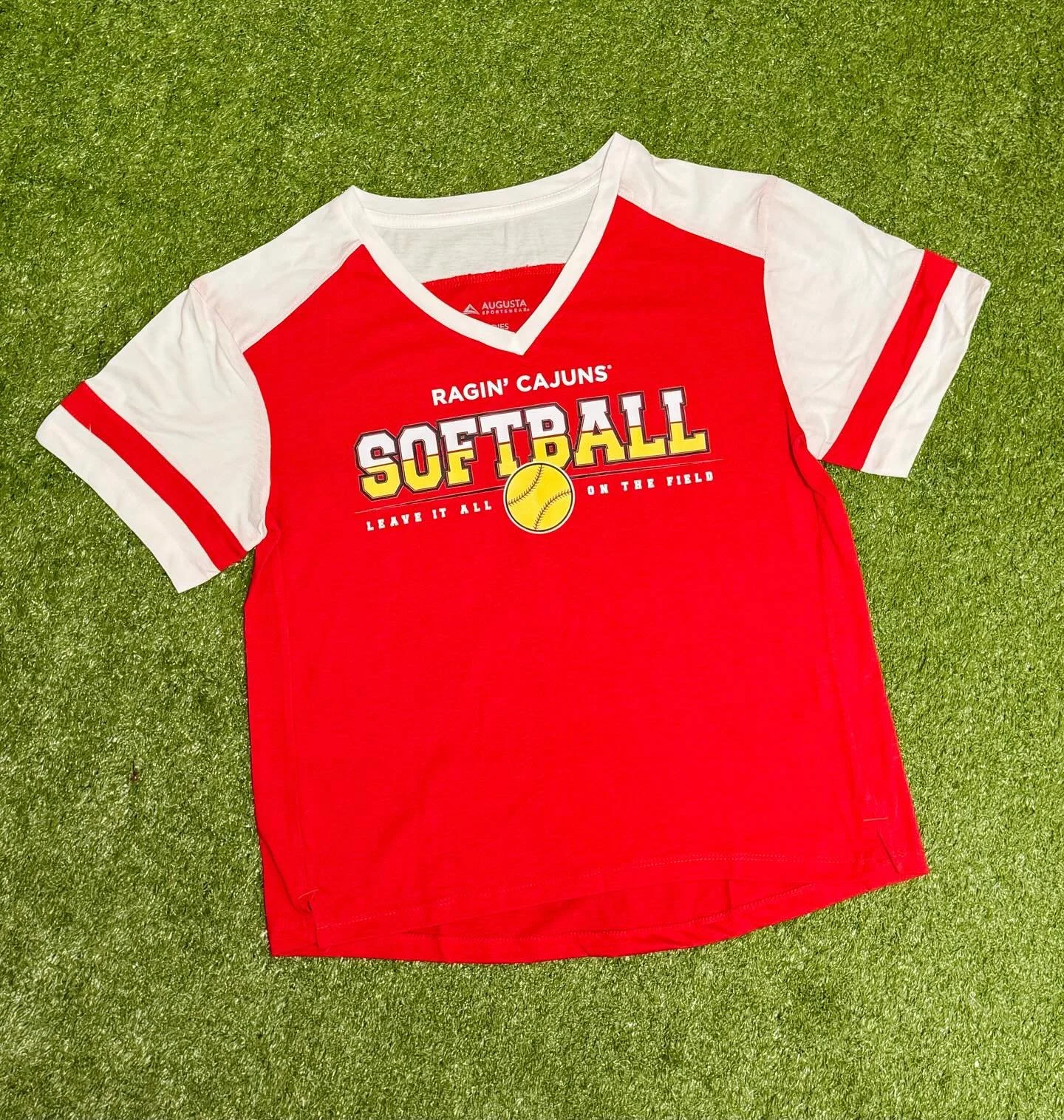Red and white softball shirt with 'Ragin' Cajuns Softball' logo, a yellow softball graphic, and the text 'Leave it all on the field,' laid on green artificial turf.