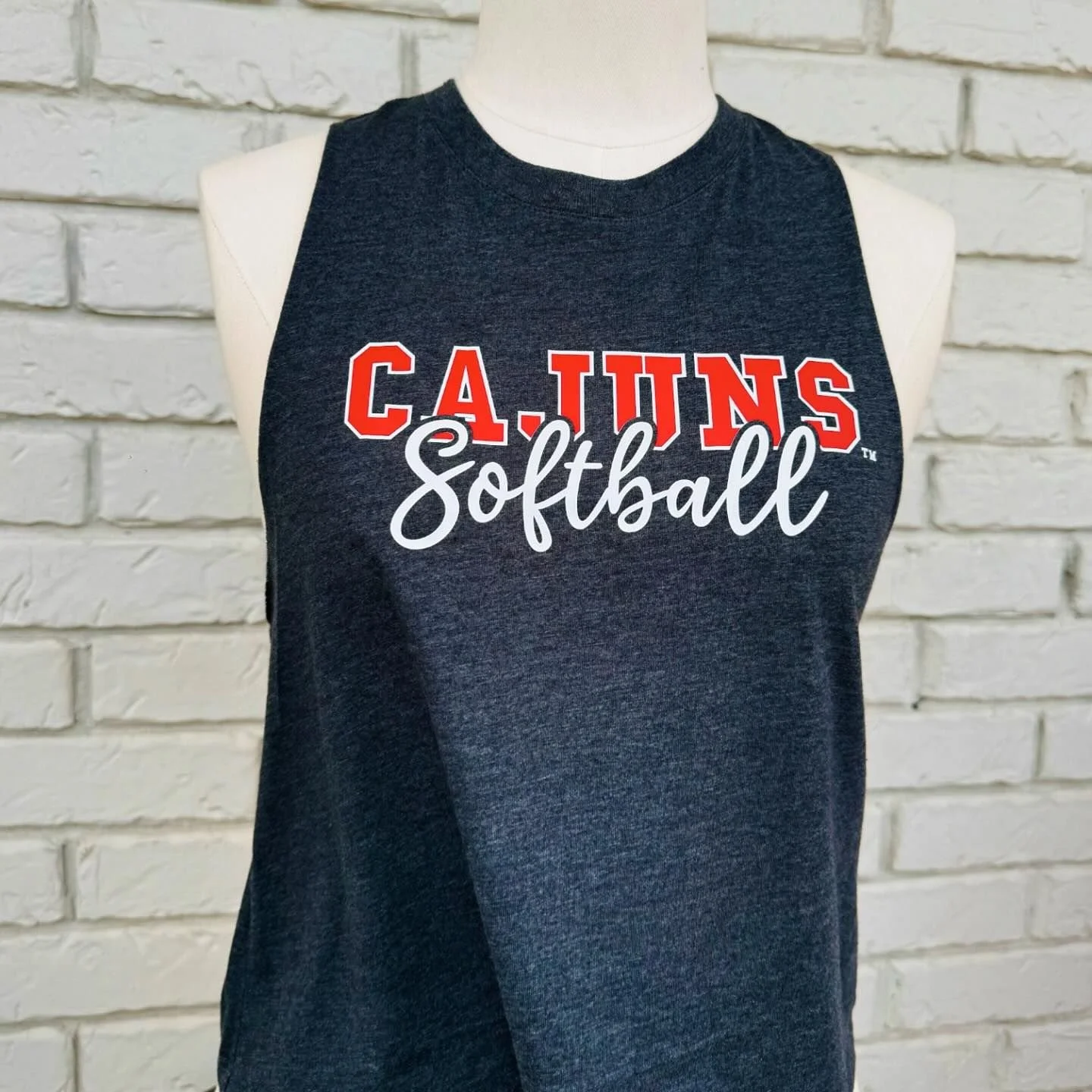 Black sleeveless shirt with red and white lettering reading "Cajuns Softball" on a white brick wall background.
