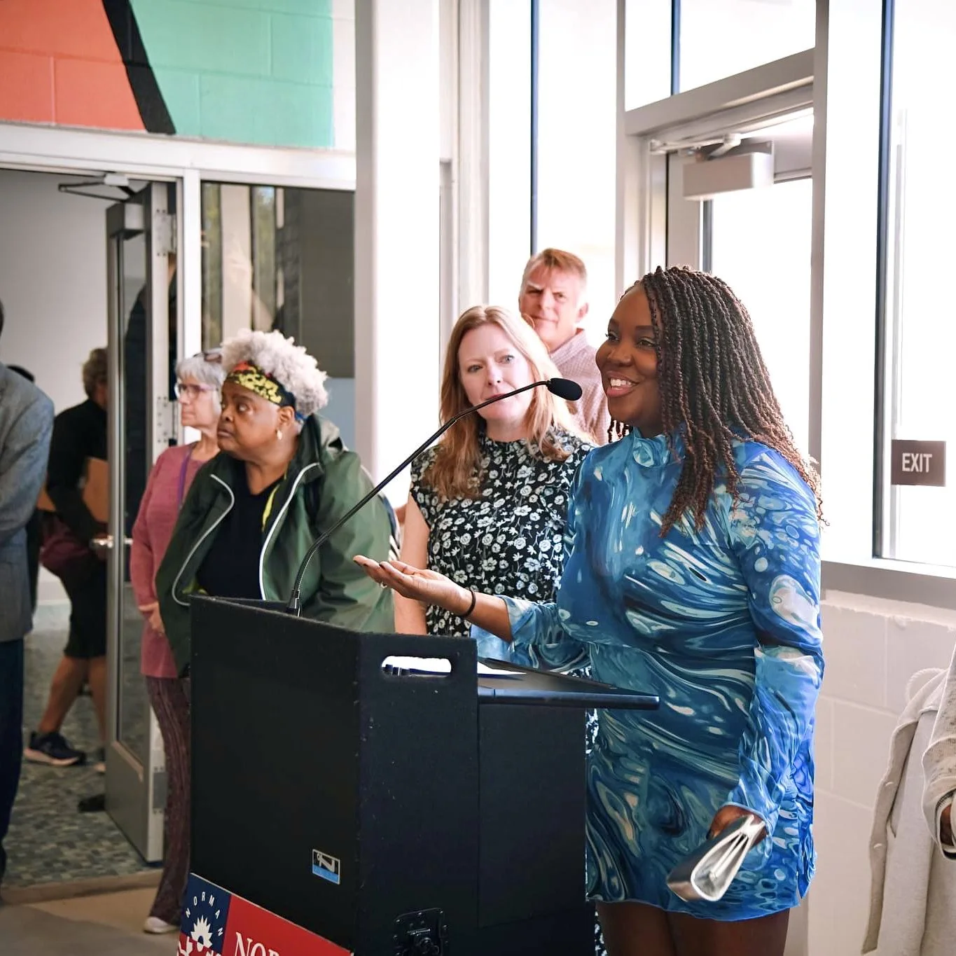 A woman in a blue dress speaking at a podium indoors with an audience behind her, by a window and exit sign.