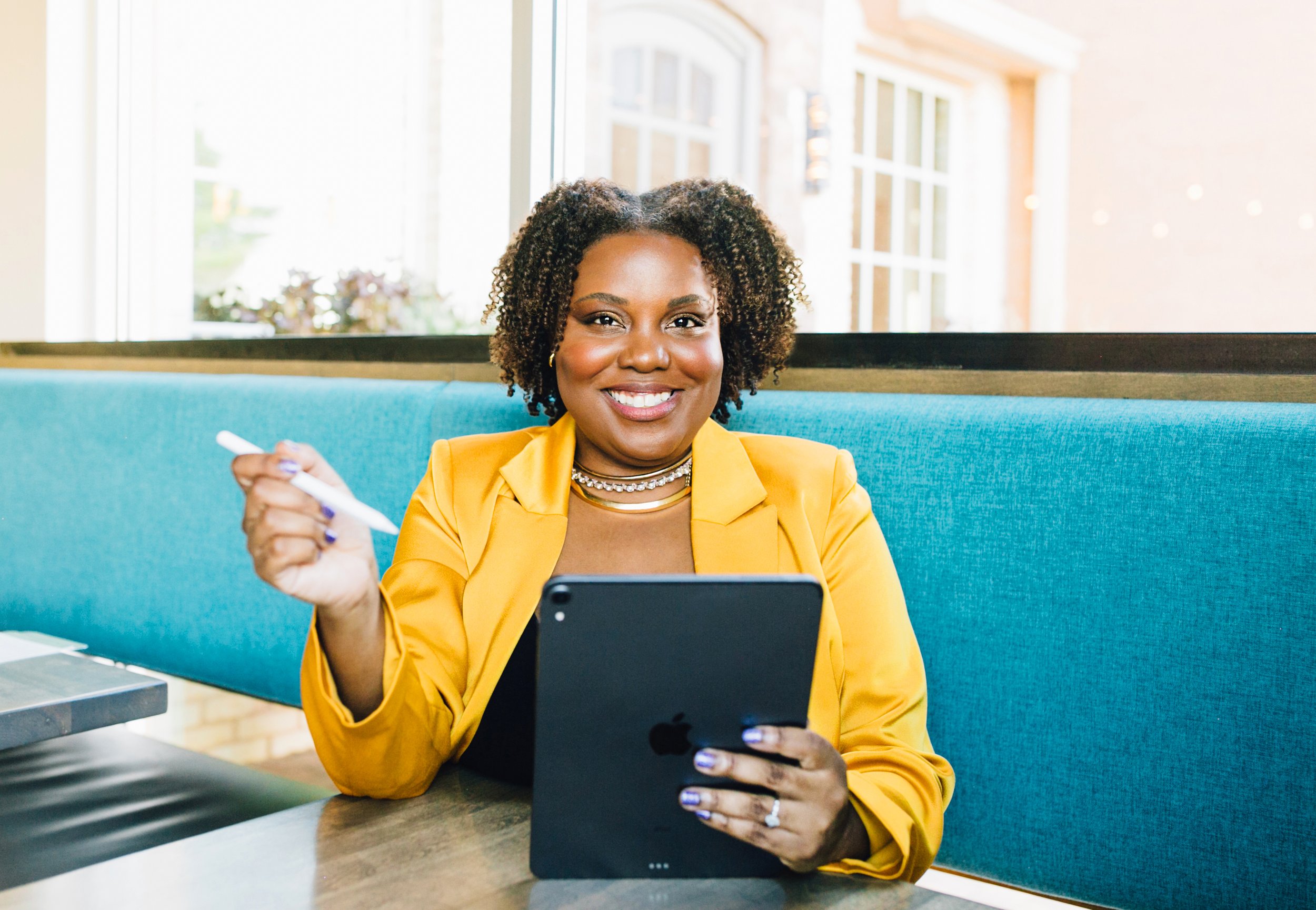 Smiling person in a yellow blazer holding a pen and tablet in a bright setting.