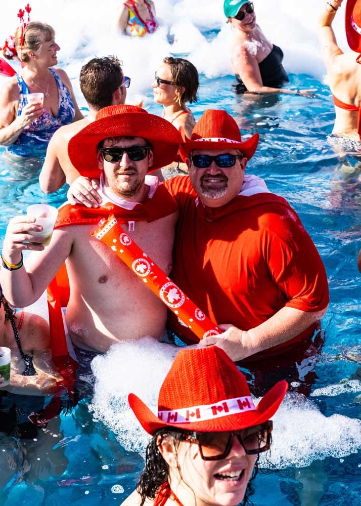 People having fun at a pool party wearing red hats and sunglasses, with foam in the water, celebrating a Canadian-themed event.