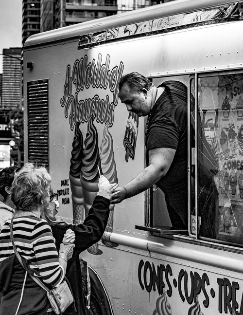 Black and white photo of a man handing an ice cream cone to two women at an ice cream truck.