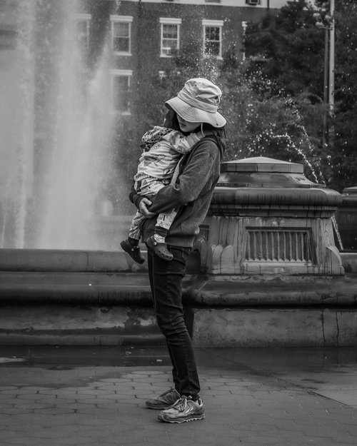 Person holding a child near a fountain, both wearing hats, in black and white photo.