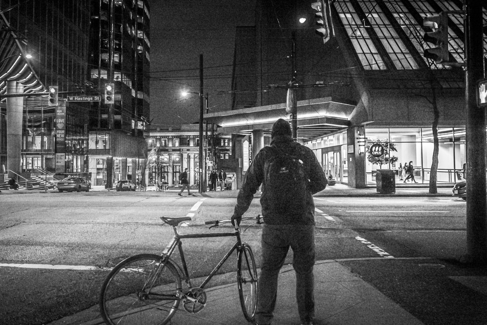 Person with bicycle at night on city street corner, surrounded by tall buildings and streetlights in black and white