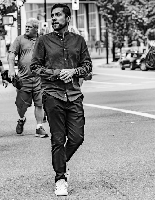 Black and white photo of a man walking confidently across a city street, dressed in a button-up shirt and pants, wearing sneakers. Several other people and cars in the background.