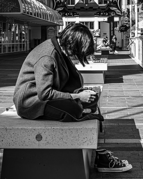 Monochrome image of a person sitting on a bench, wearing a coat, with head down, and a pair of sneakers placed beside the bench. The background features an arcade with architectural details.