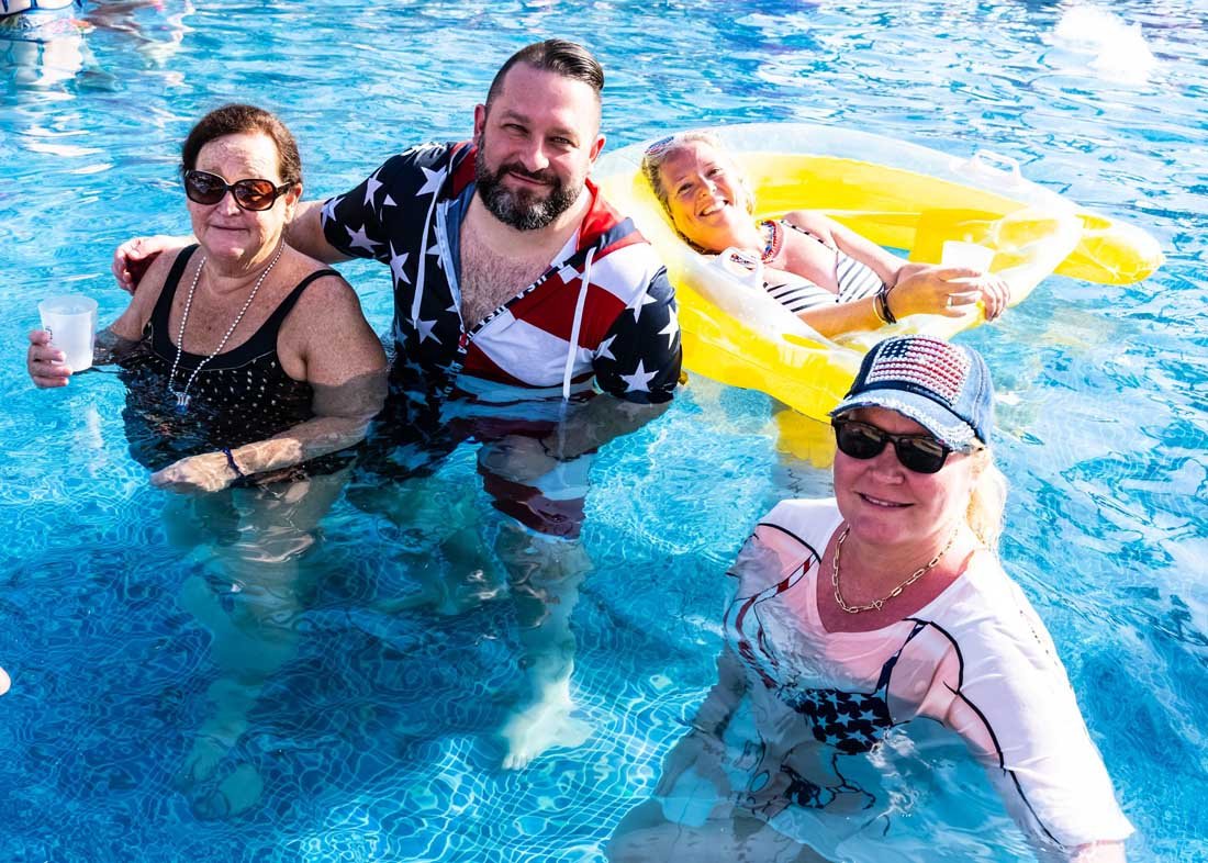 Four adults enjoying a pool. Two women wearing sunglasses and a man in a star-spangled outfit stand in the water, while another woman relaxes on a yellow inflatable raft.