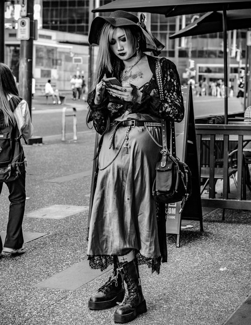 Black and white photo of a person with alternative fashion, standing on a city street. They are wearing a long skirt, lace top, platform boots, and a wide-brimmed hat. Holding a smartphone, they appear focused while pedestrians and city structures ar