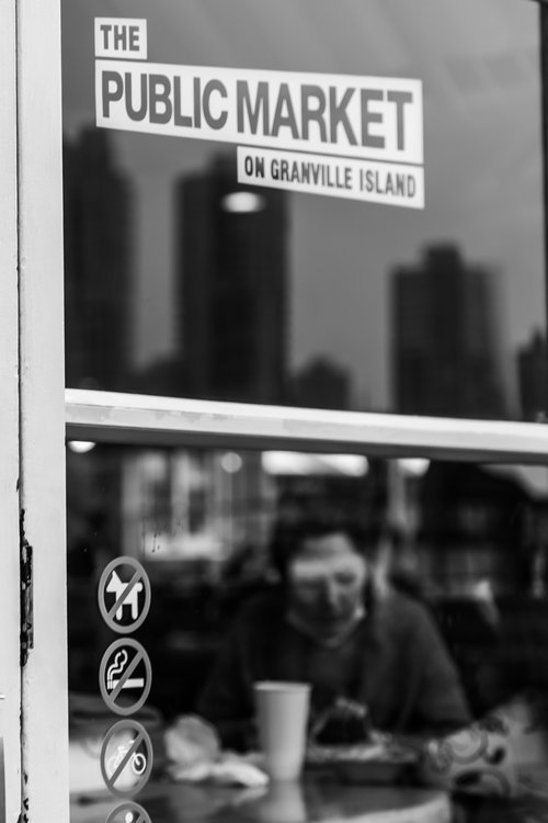 Black and white photo of a glass door with a sign reading "The Public Market on Granville Island" and various prohibition symbols. A person is visible inside, seated at a table with a drink.