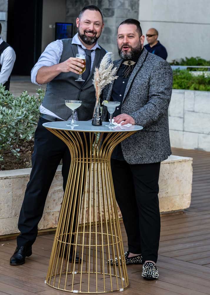 Two men standing by a high table with drinks, smiling, one holding a beer and the other with a cocktail, on an outdoor patio.